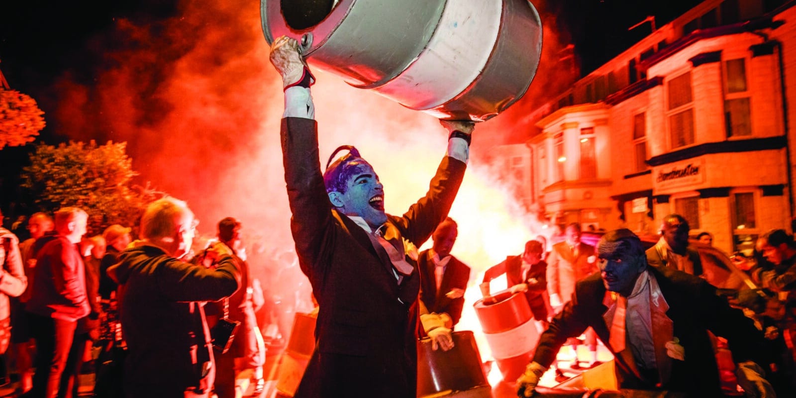 Man painted in blue holding a oil barrel above his head. He is screaming in excitement . Surrounded by other people painted in blue. Strong orange hues in the photo