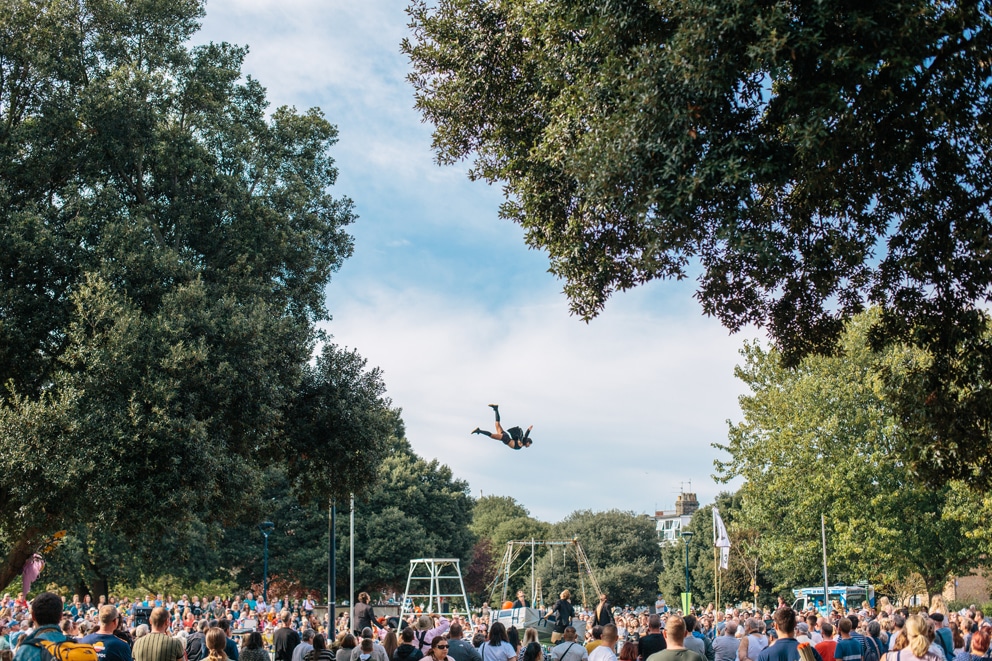 Person flying through the air from a trapeze performance. Surrounded by blue skies, trees and a big crowd.
