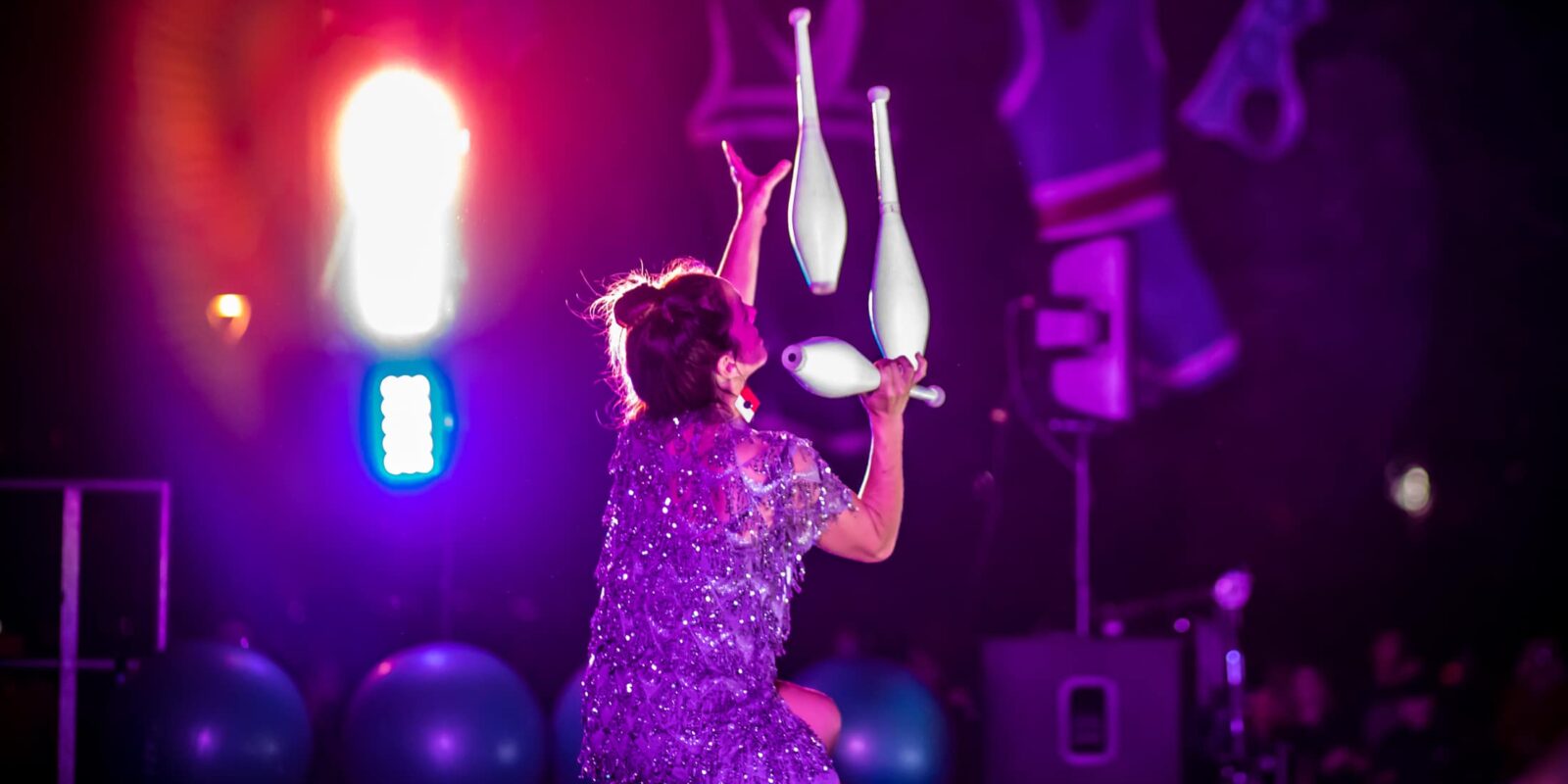 Circus Performer juggling three juggling clubs. They are balancing the clubs on each other. They are surrounded by a purple light.