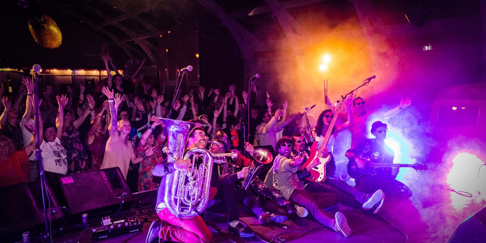 Kumpania Algazarra at Festa Fiesta 2019. A band poses for a group photo on stage during a music performance. They are carrying brass instruments and are washed in a purple light. A lively and energetic spectacle