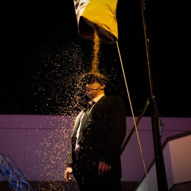 Person in suit and bowler hat standing below a bag of grain that is slowly pouring on them