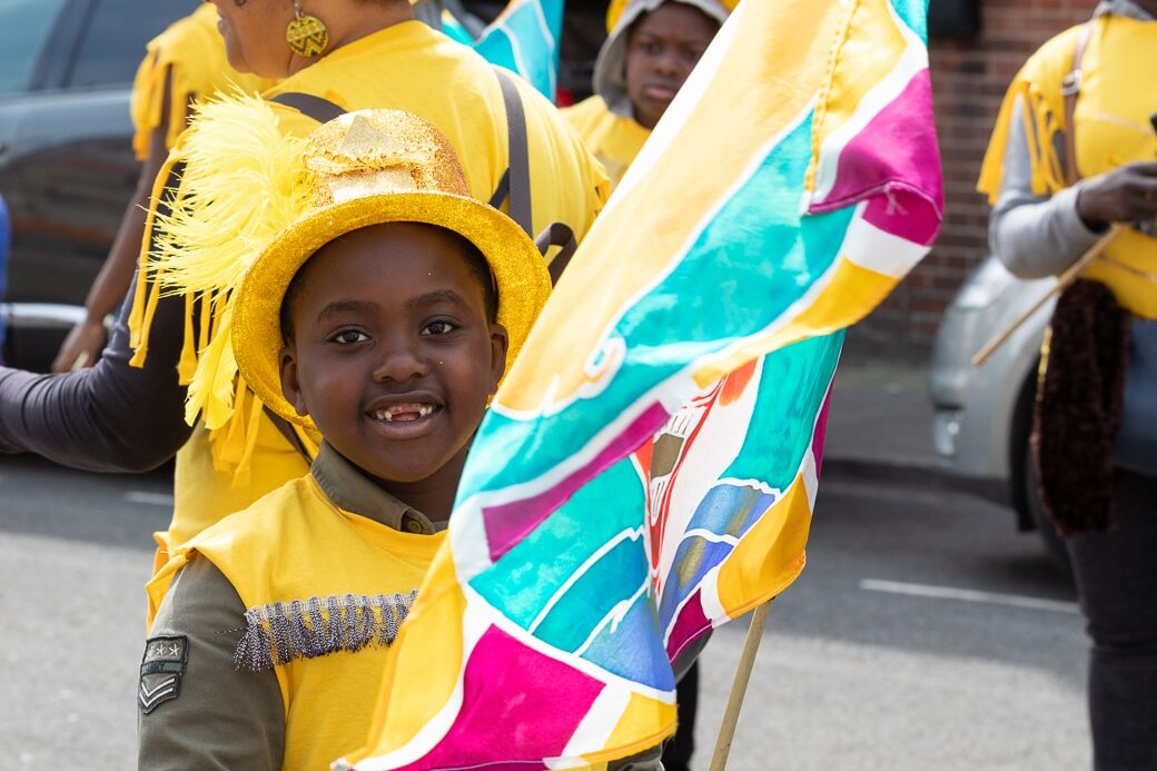 A child in bright yellow smiles at the camera while waving a flag at the Drill House