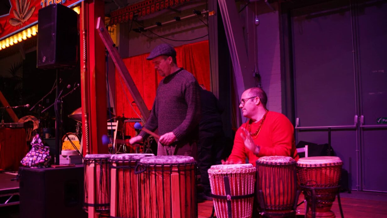 Two men play drums, with one sitting and one standing, at the Drill House