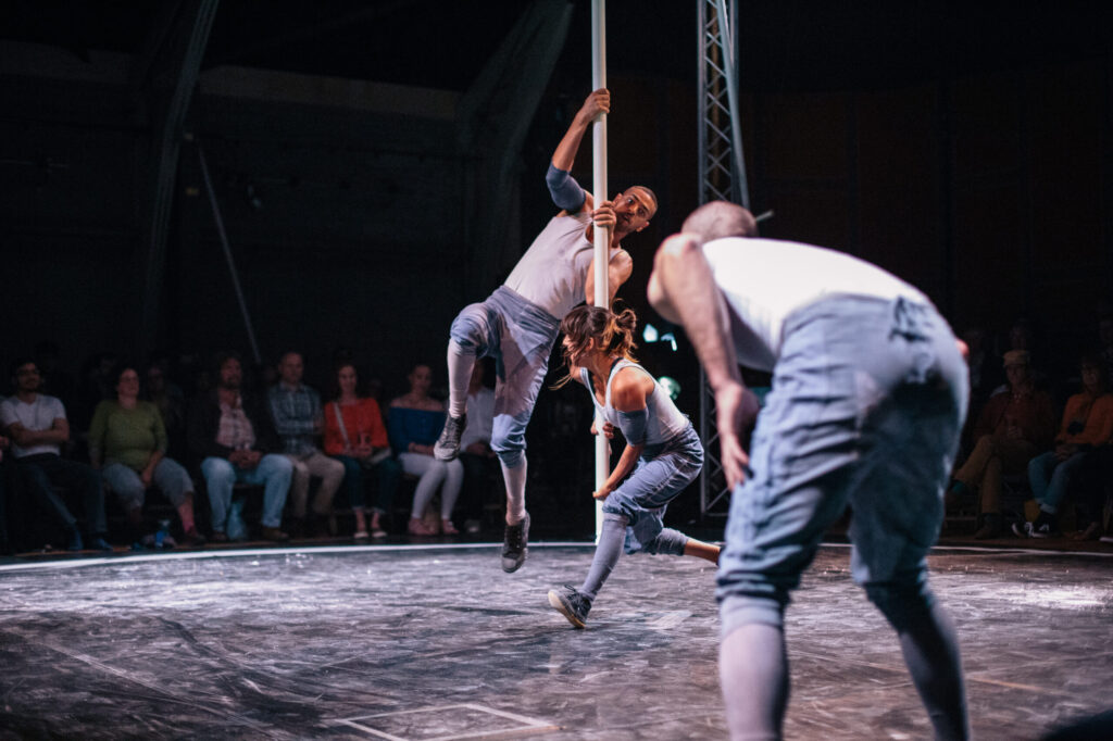 Artists perform on a stage with one in the act of swinging on a metal pole