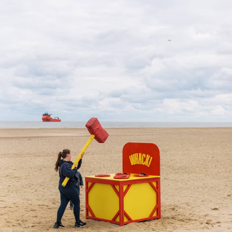 A blue sky scene on the beach. On the sand is a person playing a giant whack a mole game that is painted yellow and red.