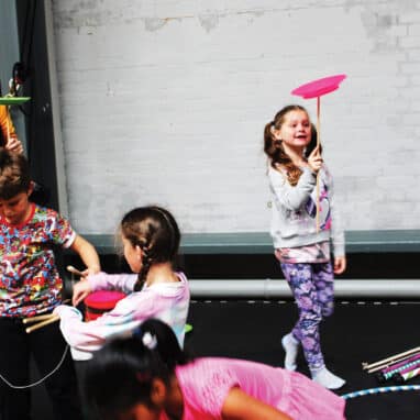 A group of children performing circus tricks while an instructor watches, at the Drill House