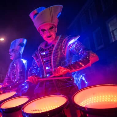 Drummers in white makeup and costume with drums lit by LED lights