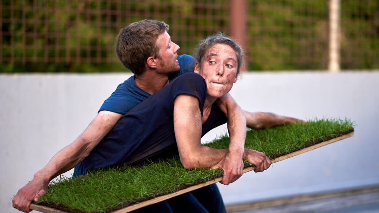 Performance of Grasshoppers. Two performers holding a plank of wood with turf grass laid on top. They contorted around each other