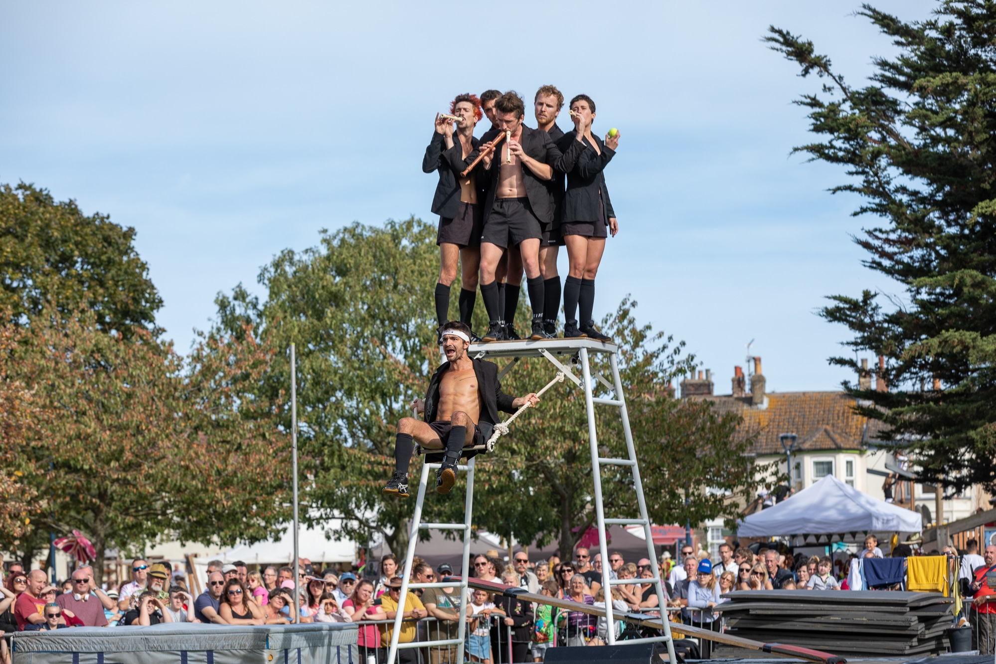 5 people stand on top a structure while a person swings below them. They are all trying to play the recorder through their nose. Surrounded by a large crowd at Out There Festival