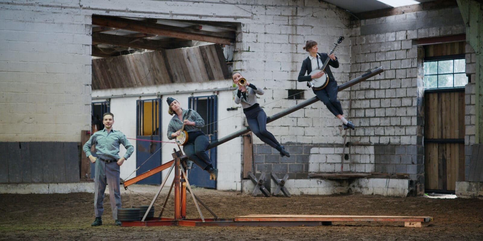 4 performers sit on a circus teeterboard all with musical brass instruments