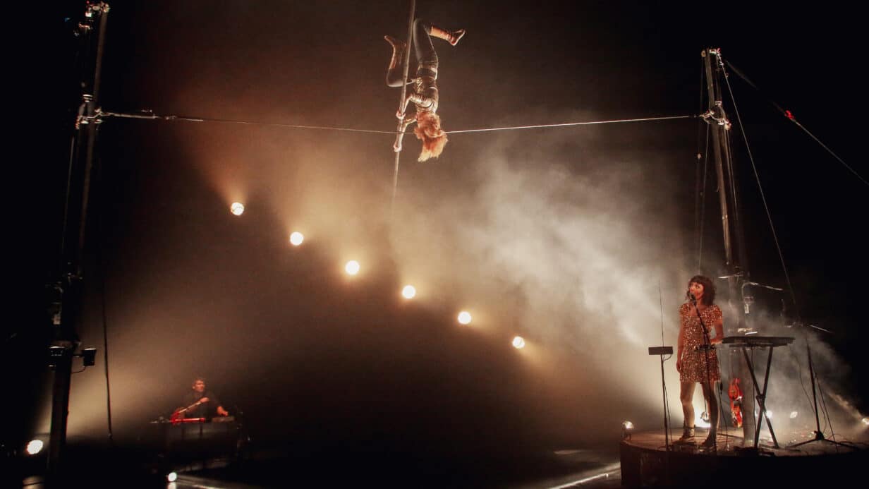 Person performing on a tightwire upside. The scene is dark with a advanced lighting display illuminating the stage. A singer performs underneath the tightwire