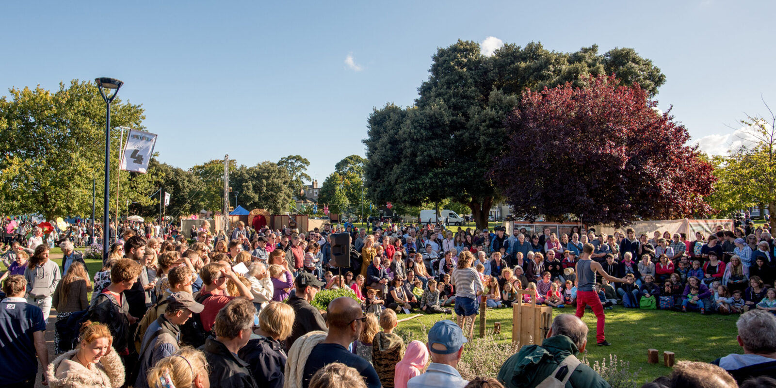 Large crowds gather in a park on a sunny day during the Out There Festival. They surround an outdoor arts performance of duo acrobatics