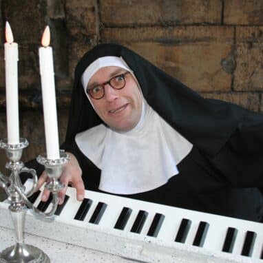 Matthew Hunt – Musical Ruth 2 – Jo Burgess a whacky nun wearing glasses playing a white piano, with a 3-sticked candle holder on top with 3 lit candles