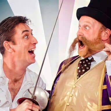 A man in a white shirt holding a fencing sword, playfully threatening a moustachioed man in a top hat