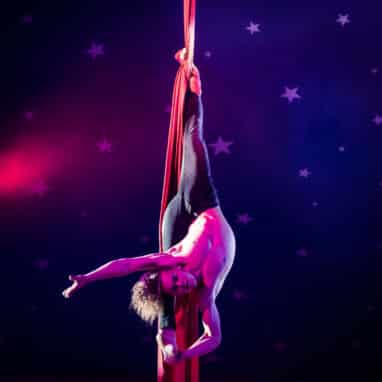 A male circus performer in leggings and a bare chest hangs upside down in the splits with one foot entwined in a piece of long fabric