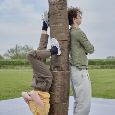 Two performers standing next to a post with their arms crossed, with one standing on their head