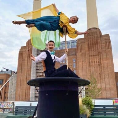 A male magician sits on top of a giant top hat with a female suspended above him leaning only on a broom, in front of Battersea Power station