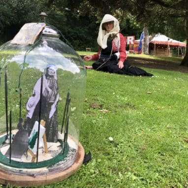 A Bell Jar in the foreground with a photograph of a Victorian woman and a woman dressed in a period-style black dress and white hat and scarf