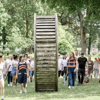 An audience and performers follow a large wooden wheel