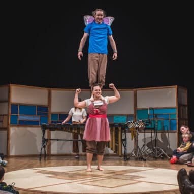 A performer in a dress is on a stage, while another performer wearing fairy wings stands on their shoulders