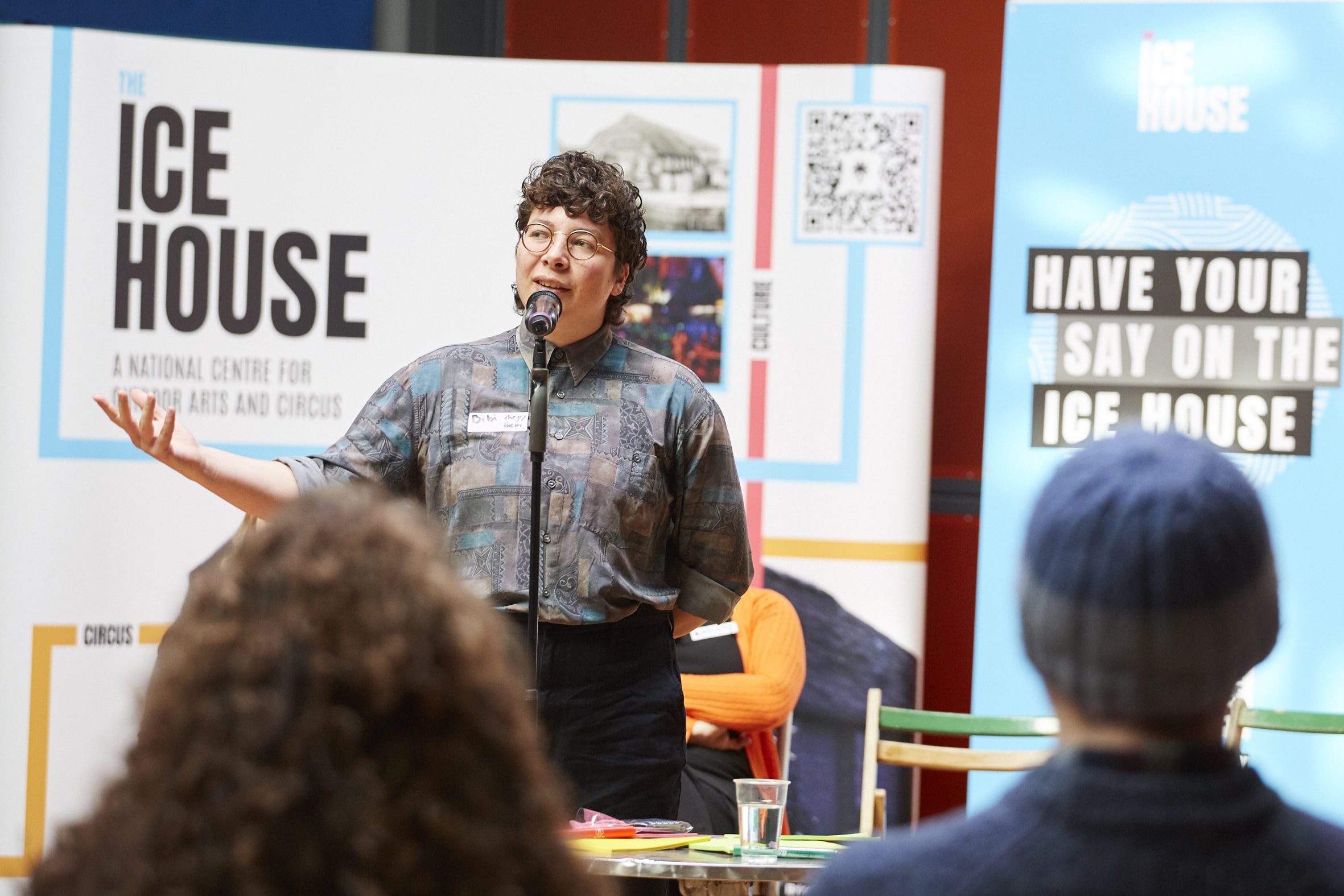 Person addressing an audience at The Drill House with The Ice House banner in the background