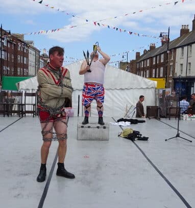 A man in chains in his underpants and another man in his underpants swallowing knives.