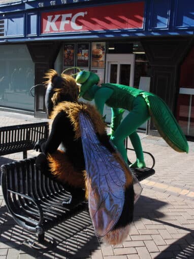Two giant insects on a part bench outside of KFC