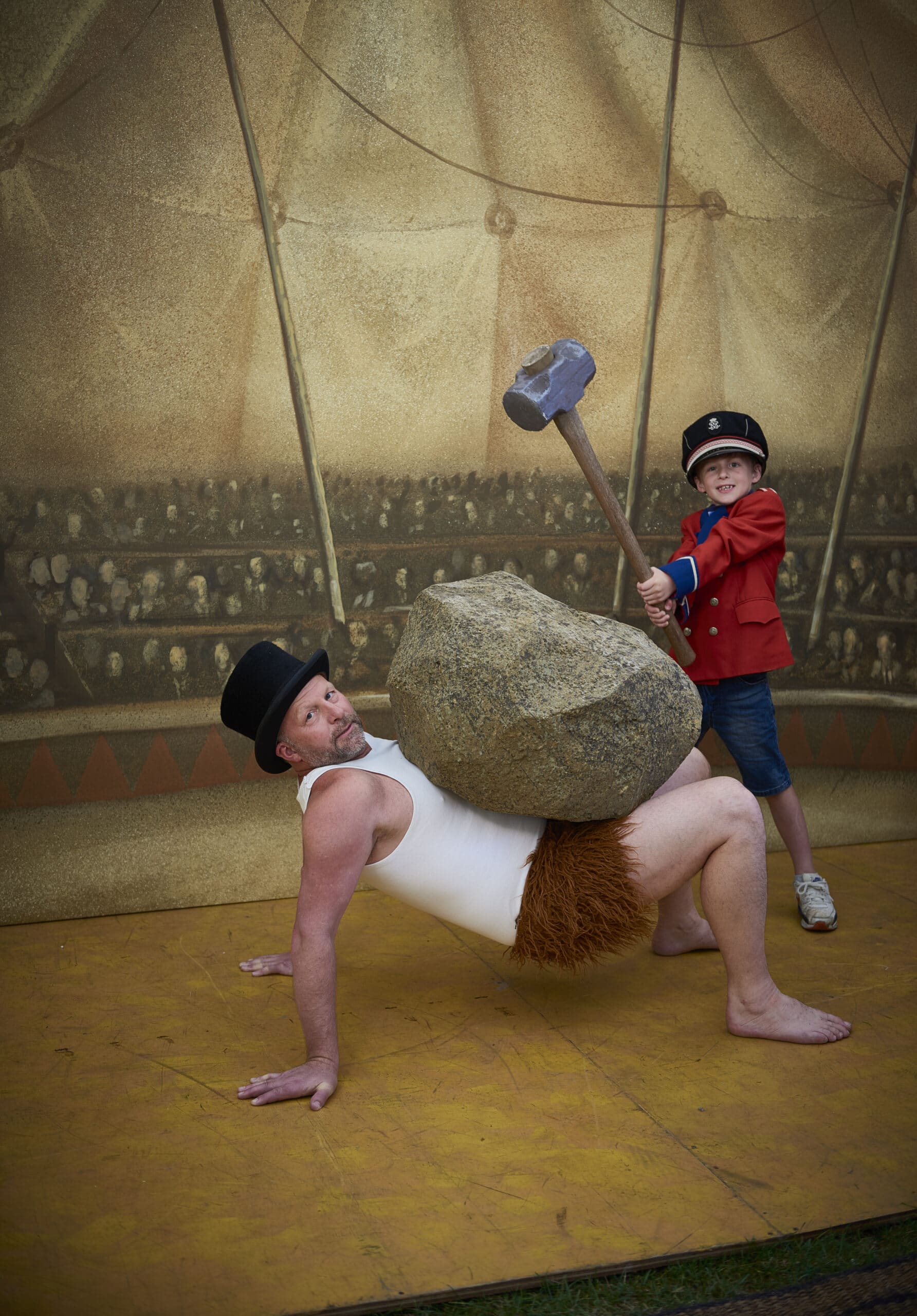 A man dressed as a strong man and a boy dressed as a ring master posing with a giant rock and hammer