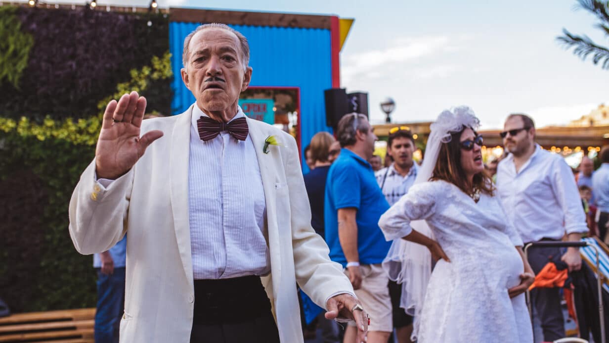 A woman dressed in a brides outfit, in tears next to a man dressed in a white tuxedo like a mafiaoso