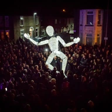 Giant illuminated puppet surrounded by a large crowd at the Out There Festival