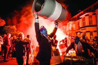 Man in Blue paint holding barrel above his head surrounded by fire at the Out There Festival